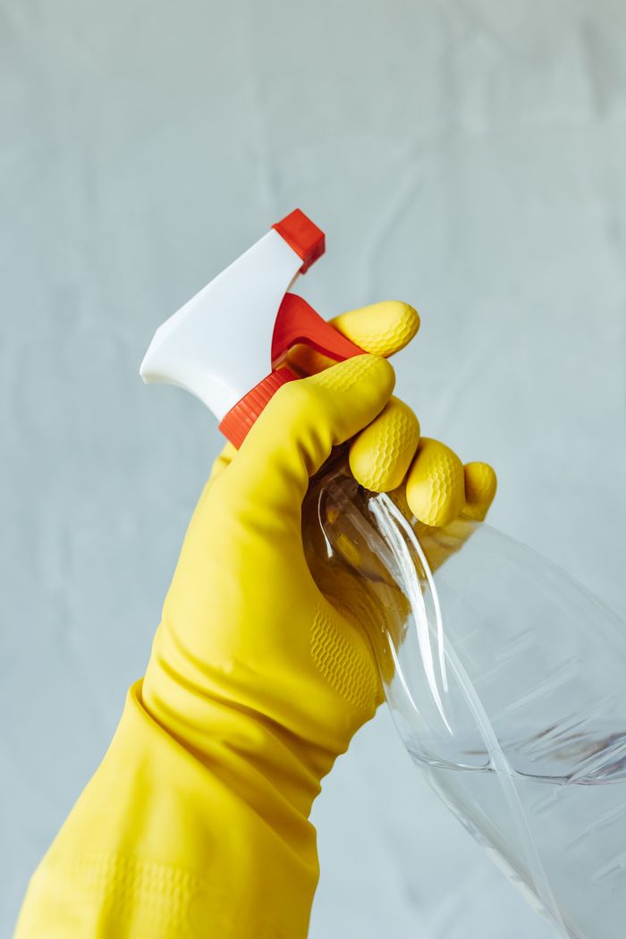 A hand in a yellow rubber glove is holding a red-and-white spray bottle in front of a plain neutral background.